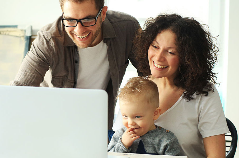 En familj på tre personer tittar in i dator och ler.